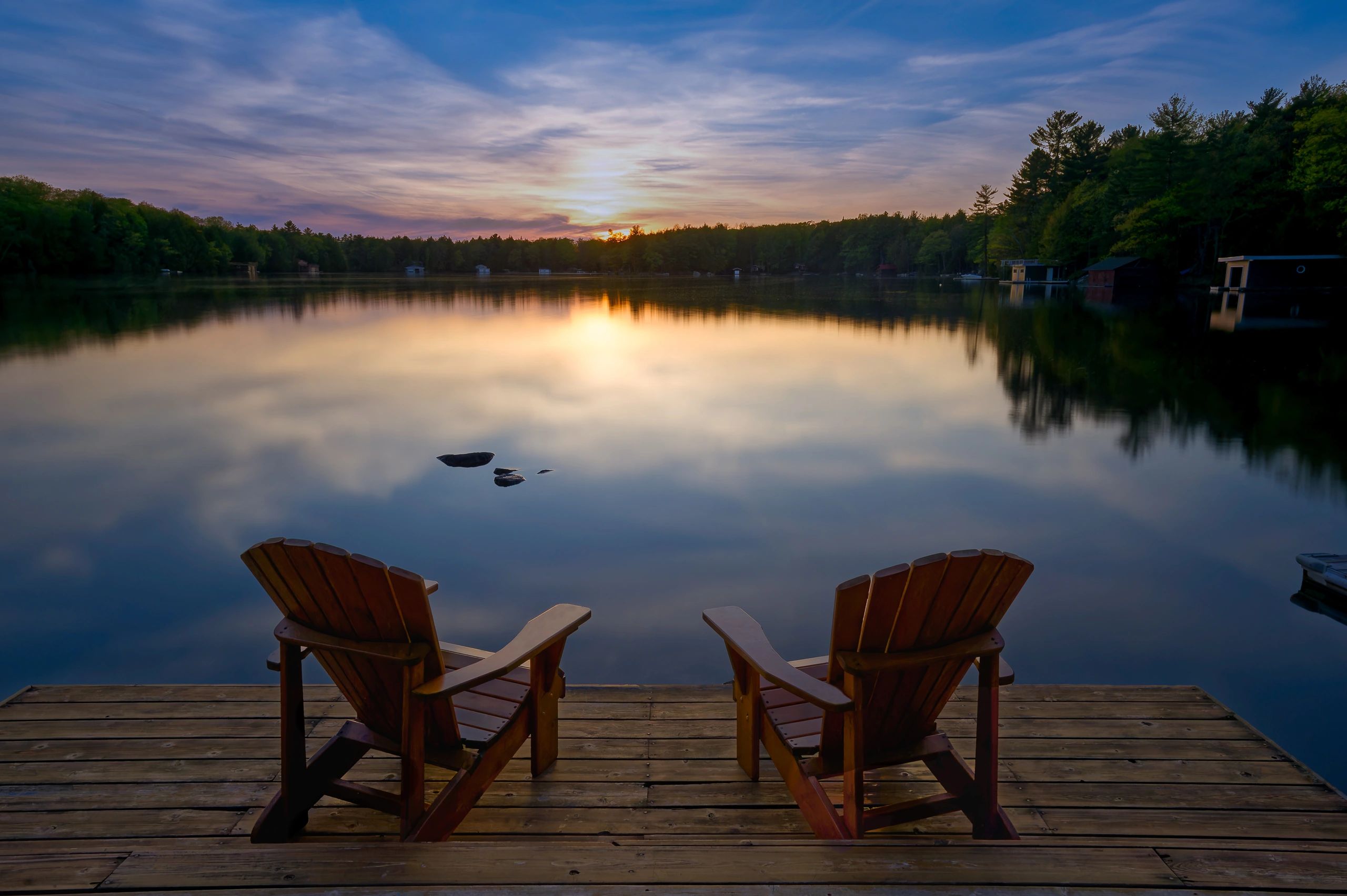 Wooden dock with Adirondack chairs overlooking a peaceful lake at sunset